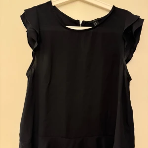Svart blus med volangärm Forever 21 - Stilren svart blus från Forever 21 med volangdetaljer på ärmarna och rund halsringning. Toppen är ärmlös och har en synlig dragkedja i ryggen.  Blusen är i jätte fint skick!