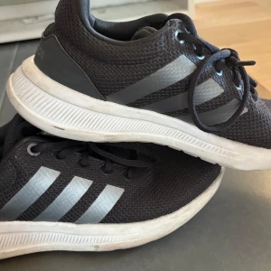 Svarta Adidas sneakers med mesh - Svarta Adidas sneakers med klassiska tre ränder i silver på sidorna. Ovandel i ventilerande mesh och snörning framtill. Vit, mönstrad sula för bra grepp. Perfekta för dig som gillar sportig och clean stil.