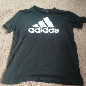 Svart Adidas t-shirt med logga - Svart t-shirt från Adidas med stor vit logga framtill. Klassisk rund hals och korta ärmar. Tillverkad i mjuk bomull som är skön att ha på sig. Perfekt för dig som gillar sportig och enkel stil.