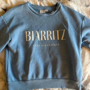 Blå sweatshirt från Gina Tricot - Snygg blå sweatshirt från Gina Tricot med trycket 'BIARRITZ CÔTE ATLANTIQUE' framtill. Tröjan har rund hals, ribbade muddar och är tillverkad i mjuk bomullsmix. Perfekt för en chill och avslappnad stil.