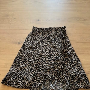 Leopardmönstrad omlottkjol Lindex - Snygg leopardmönstrad omlottkjol från Lindex med volangkant och knyt i midjan. Kjolen har beige botten med svarta och bruna fläckar, vilket ger en trendig look. Perfekt för dig som vill sticka ut lite extra.