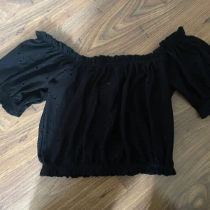 Svart offshoulder topp Gina Tricot XS - Supersöt svart offshoulder topp från Gina Tricot i storlek XS. Toppen har broderade hål och resår vid axlar och nederkant för en snygg passform. Perfekt till jeans eller kjol när du vill ha en trendig look.