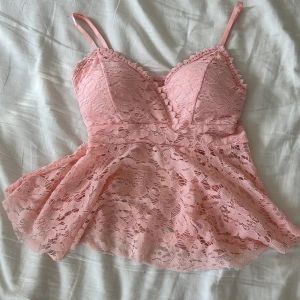 Rosa spetsig linnetopp med volang - Supersöt rosa linnetopp i spets med smala axelband och v-ringad byst. Toppen har volang nertill och fina spetsdetaljer längs kanterna. Går att justera banden och är väldigt stretchig 