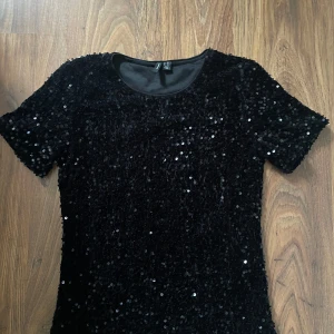 Svart paljettopp från Vero Moda - Glittra loss i denna svarta topp från Vero Moda, täckt med paljetter som fångar ljuset. Modellen har rund halsringning och korta ärmar, perfekt för att sticka ut. Snygg och trendig att styla med jeans eller kjol för en cool look. Använd endast 3 gånger
