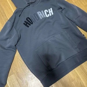 Grå HOODRICH hoodie med brodyr - Snygg grå hoodie från HOODRICH med broderad logga i svart, grått och vitt på bröstet. Klassisk känguruficka framtill, huva med dragsko och mjukt material som känns skönt mot huden. Perfekt för en chill och stilren streetlook.