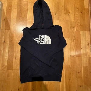 Mörkblå hoodie från The North Face - Snygg mörkblå hoodie i storlek xl (barnstorlek) från The North Face med stor vit logga på bröstet. Klassisk känguruficka framtill och huva. Perfekt för dig som gillar streetwear och vill ha en bekväm och stilren tröja.
