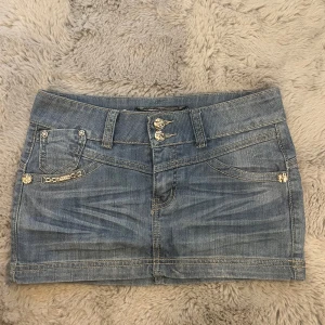 Söt mini kjol - Snygg ljusblå jeanskjol med coola silvriga knappar och blingdetaljer på fickorna. Kjolen har klassiska jeanssömmar, dekorativa nitar och en liten kedja framtill. Perfekt för dig som gillar en trendig och avslappnad stil. Perfekt mini kjol!