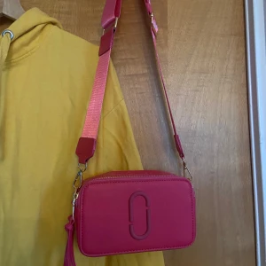 Rosa axelväska från Marc Jacobs - Säljer en rosa axelväska med bred axelrem i textil och logga. Väskan är i skinn med två dragkedjor upptill och har en stilren, boxig form. Köpt här på plick av en säljare.