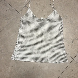 Vit prickig linnetopp från H&M XS - Supersöt vit linnetopp från H&M i storlek XS med tunna axelband och små svarta prickar över hela tyget. Toppen har en lätt och luftig känsla, perfekt för varma dagar. Enkel och stilren design som är lätt att matcha med jeans eller kjol.