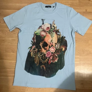 Ljusblå McQueen t-shirt - Ljusblå t-shirt från McQueen med stor konstnärlig dödskalle- och blomprint på framsidan. Baksidan har vit McQueen-logga. Klassisk passform och korta ärmar, tillverkad i mjuk bomull. Perfekt för dig som gillar edgy och unik stil.
