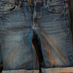 Blå jeans Shorty Star stl 150 - Snygga blå jeans från Shorty Star i storlek 150. Klassisk femficksmodell med uppvikta benslut och slitningar för en cool look. Jeansen har normal passform och är tillverkade i mjukt denimtyg med dragkedja och knapp framtill.