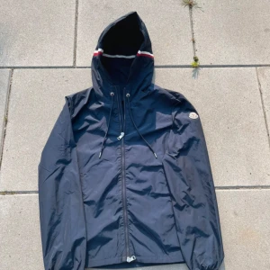 Mörkblå vindjacka från Moncler - Snygg mörkblå vindjacka från Moncler med huva och dragsko. Jackan har dragkedja framtill, Moncler-logga på ärmen och en vit/röd detalj på huvan. Perfekt för dig som vill ha en stilren och sportig look.