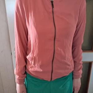 Rosa bomberjacka med dragkedja - Säljer en stilren rosa bomberjacka med hel dragkedja framtill. Jackan har långa ärmar, ribbade muddar och en klassisk bomberpassform. Perfekt för dig som vill ha en enkel men snygg jacka till vardags.