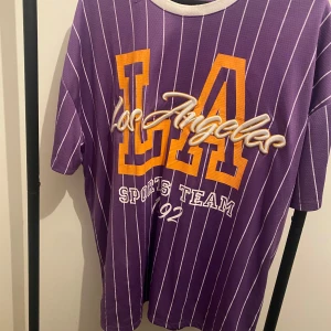 Lila randig t-shirt LA FB Sister XL - Lila t-shirt från FB Sister i storlek XL med vita vertikala ränder och stor LA-logga i gult och vitt framtill. Sportig vibe med vit ribbad krage och tryck 'Los Angeles Sports Team 92'. Oversized passform och mjukt polyestertyg.