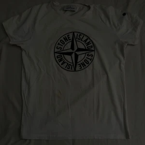 Vit Stone Island t-shirt med logga - Vit t-shirt från Stone Island med stor svart kompasslogga på bröstet. Klassisk passform och korta ärmar. Tillverkad i mjuk bomull och har svarta knappar på ärmen som snygg detalj. Perfekt för dig som gillar streetwear och vill sticka ut. DU KAN SÄTTA FAST DITT STONE ISLAND MÄRKE PÅ AXELN