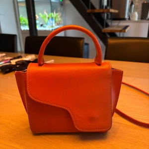 Orange handväska från ATP Atelier - Snygg orange handväska från ATP Atelier i slätt skinn. Väskan har ett unikt lock med asymmetrisk form, ett kort handtag och en lång axelrem som går att ta av. Stängs med magnetknapp och har stilrena metalldetaljer. Perfekt för dig som vill sticka ut med färg. !OBS! Väskan har defekter och är väl använd. Det är en hel del repor och har tappat färg som visas på bilderna. Är även köpt här på Plick så har varken kvitto, kartong eller dustbag till. Disk. pris vid snabb affär!