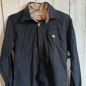 Svart Burberry skjorta  - Svart skjorta från Burberry med klassiska rutiga detaljer på insidan av kragen och ärmsluten. Jättesnygg och stilren. Storlek S, men något liten i storleken. Säljer för att jag inte fått någon användning av den. Den är något skrynklig på bilderna, men jag kan stryka den innan jag skickar iväg den. ☺️