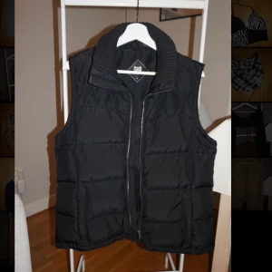 Svart dunväst från Flash med krage - Svart dunväst från Flash med bred, ribbstickad krage och dragkedja framtill. Västen är quiltad och har en klassisk, rak passform. Perfekt att slänga över en hoodie för extra värme. Snygg och enkel design som funkar till det mesta.