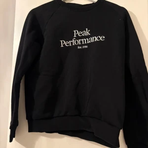 Svart Peak Performance sweatshirt - Svart sweatshirt från Peak Performance med vit broderad logga på bröstet. Klassisk rund halsringning och långa ärmar. Mjuk och skön bomullsblandning, perfekt för en chill och stilren look.