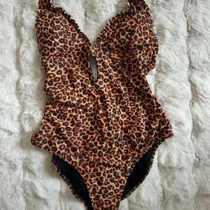 Leopardmönstrad baddräkt från Nelly - Snygg baddräkt från Nelly med leopardmönster i bruna och svarta toner. Djup urringning med volangkant och cut-out framtill. Justerbara axelband och fodrad insida. Tillverkad i stretchig polyesterblandning för perfekt passform.