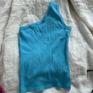 Turkos oneshoulder topp från Stockholm  - Snygg turkos ribbad topp med oneshoulder-design. Perfekt för dig som vill sticka ut med en cool och modern look. Toppens material är stretchigt och sitter skönt mot kroppen. Passar perfekt till jeans eller kjol för en trendig outfit.
