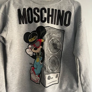 Grå Moschino x H&M tröja med Mickey - Grå sweatshirt från H&M x Moschino med stor svart logga och coolt tryck av Mickey Mouse i streetstyle bredvid en högtalare. Tröjan har rund hals, långa ärmar och är gjord i mjuk bomullsmix. Perfekt statement piece för dig som gillar unika prints.