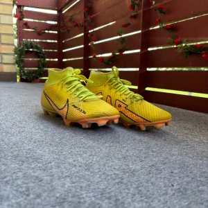 Nike Mercurial Air Zoom Academy fotbollskor - Nike Mercurial Air Zoom fotbollsskor storlek 44,5  i en riktigt fräsch gul färg med orange detaljer och svart swoosh. Skorna har snörning, högre strumpliknande skaft och mönstrad sula för grymt grepp på planen. Perfekta för dig som vill sticka ut och ha max kontroll på bollen. Har användning i 3-5 månader.