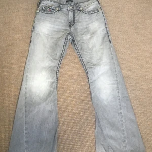 True Religion Joey Low Rise Flare Jeans - Snygga grå jeans från True Religion, modell Joey Low Rise Flare. Byxorna har låg midja, utsvängda ben och coola kontrastsömmar. Bakfickorna har den klassiska True Religion-bågen i rött. Perfekta för dig som gillar en unik och avslappnad stil.