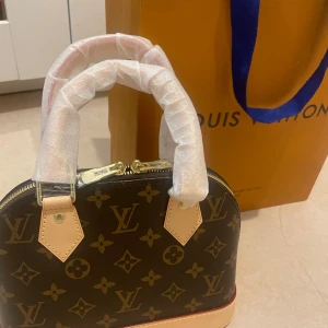 Louis Vuitton Monogram handväska - Säljer en ikonisk Louis Vuitton handväska i klassiskt monogrammönster. Väskan är brun med beiga detaljer och har guldfärgade dragkedjor. Tillverkad i skinn med LV-loggor och blommor över hela väskan. Kommer med dustbag och originalförpackning.