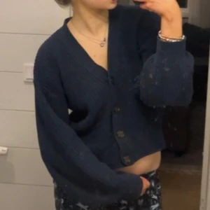 Mörkblå stickad kofta från H&M - Mysig mörkblå stickad kofta från H&M i storlek XS. Koftan har ribbade detaljer, v-ringning och knäpps med fyra knappar framtill. Perfekt att slänga över en t-shirt eller linne när det är kyligt ute.