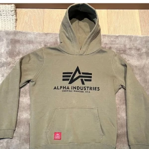 Alpha Industries Hoodie  - Säljer en olivgrön hoodie från Alpha Industries med stor logga på framsidan. Tröjan har en klassisk känguruficka och ribbade muddar vid ärmslut och nederkant. Perfekt för en avslappnad stil.