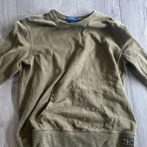Ralph lauren sweatshirt  - Olivgrön sweatshirt från Polo Ralph Lauren med klassisk rund hals och stor magficka framtill. Tröjan har långa ärmar och en avslappnad passform, perfekt för chill dagar. Materialet är mjuk bomull som känns skönt mot huden.