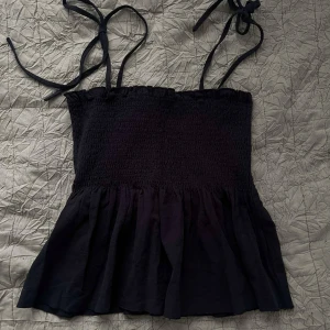 Svart linnetopp med knytband - Supersöt svart linnetopp med smockad överdel och volang nertill. Axelbanden knyts på axlarna för en personlig touch. Perfekt till jeans eller kjol när du vill ha en chill men ändå snygg look.