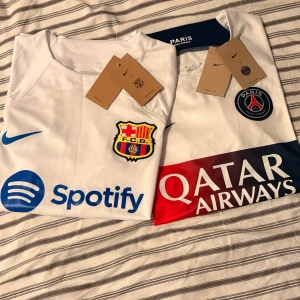Vit FC Barcelona och PSG fotbollströja Nike - Två snygga fotbollströjor från Nike: en vit FC Barcelona med blå detaljer, Spotify-logga och klubbmärke, samt en vit PSG med röd och marinblå panel, Qatar Airways-logga och PSG-emblem. Köp båda för 550 eller en för 300. 
