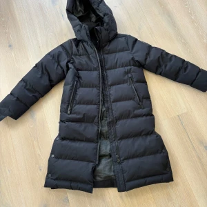 Svart lång jacka stl 146 Everest - Lång svart jacka från Everest med huva och två dragkedjeförsedda fickor. Jackan är quiltad och har en rak passform med långa ärmar. Sprund i sidan och reflexer på ärmarna. Mycket fint skick, använd en säsong.