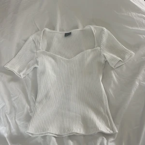 Vit ribbad topp från Gina Tricot - Superfin vit ribbad topp från Gina Tricot med korta ärmar och snygg v-ringad halsringning. Toppen har en figurnära passform och är gjord i mjukt, stretchigt material som sitter skönt. Perfekt till jeans eller kjol för en clean look.