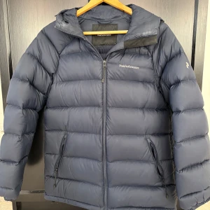 Mörkblå dunjacka Peak Performance - Snygg mörkblå dunjacka från Peak Performance med huva och dragkedja framtill. Jackan har två fickor med dragkedja och diskret logga på bröstet och ärmen. Perfekt för kalla dagar och riktigt skön att bära. Den är köpt för 3500kr. Den är i storlek M.💓💓