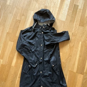 Svart regnjacka - Snygg svart regnjacka från 157 med huva och vita dragsnören. Jackan har silvriga knappar, stora fickor och är i ett galonliknande material. Jag säljer även en likadan jacka fast i mörkblått om det är av intresse. Hör av dig om du har frågor! 😊