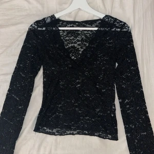 Svart spetsblus med v-ringning - Snygg svart spetsblus med v-ringning. Långärmad och figurnära passform, perfekt för att lyfta vilken outfit som helst. Blusen har transparent känsla och fina spetsdetaljer över hela plagget.