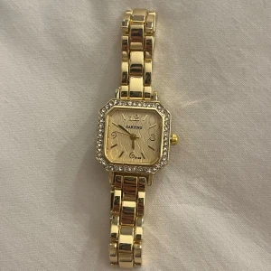 Liten guldig klocka med stenar - Snygg guldig klocka med fyrkantig urtavla, glittrande stenar runt boetten och vågigt mönster på urtavlan. Eftersom jag inte använder guld så har denna aldrig kommit till användning därför den är i helt nytt skick