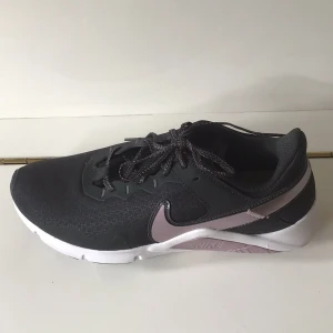 Svarta Nike sneakers med lila swoosh - Snygga svarta Nike Legend Essential 2 training shoes med ljuslila swoosh och detaljer. Ovandel i textil och syntet, vit och svart sula med mönstrad undersida. Skorna har snörning och en sportig vibe. Nya men utan prislapp.