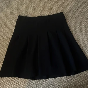 Svart plisserad kortkjol - En svart kortkjol med plisserade veck och medel midja. Kjolen har en klassisk A-linjeform som ger en snygg siluett. Perfekt att styla med en stickad tröja eller crop top. Materialet känns mjukt och följsamt. I resår tyg