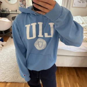 Blå Champion LIU hoodie med tryck - Säljer en ljusblå hoodie från Champion med stort vitt LIU-tryck och universitetslogga på bröstet. Tröjan har huva med snörning, magficka och ribbade muddar.🥰