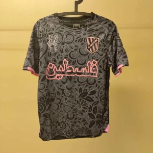 FC Palestina grå fotbollströja - Unik fotbollströja från FC Palestina i grått med svart blommigt mönster och rosa detaljer. Kortärmad modell med klubbmärke på bröstet och tryck i rosa arabiska bokstäver. Tillverkad i lätt och ventilerande funktionsmaterial.HELT NYTT OCH ÄLKTA