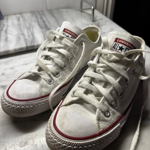 Vita Converse All Star sneakers - Klassiska vita Converse All Star sneakers med lågt skaft, röd och blå rand längs sulan och vit gummitå. Skorna har snörning, canvasmaterial och är i superfint skick!