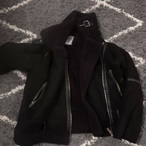 Svart teddyjacka från Stradivarius - Säljer en svart teddyjacka från Stradivarius med mysigt foder, dragkedjor på sidorna och coolt spänne vid kragen. Jackan har en oversized vibe och detaljer i skinnimitation. Perfekt för kalla dagar när du vill vara både varm och snygg.