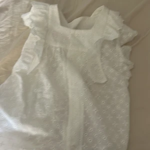 Vit blus med volang från Mini Malina - Superfin vit blus från Mini Malina med broderade detaljer och volang vid axlarna. Blusen är ärmlös och har en söt, romantisk känsla med broderad spets och vågig kant nedtill. Perfekt för dig som gillar en feminin och somrig stil. blusen kan passa till elva åringar tioåringar och nioåringar också