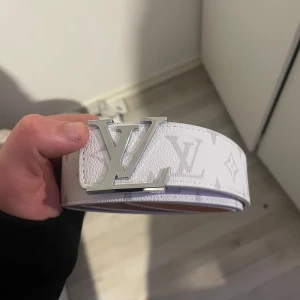 Vit  skärp med silverdetaljer  - Säljer ett vitt bälte  i skinn med klassiskt monogrammönster och stor -logga i silver framtill. Bältet har exklusiv känsla med präglad insida och tydliga detaljer. Perfekt accessoar för att lyfta din outfit. Man får me så man kan göra större hål 