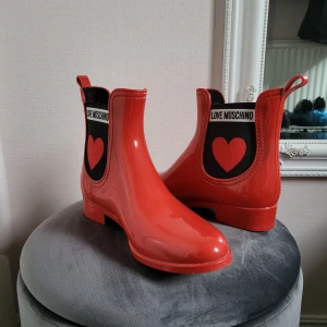 Röda boots från Love Moschino - Snygga röda boots från Love Moschino i glansig syntet med svart detalj och rött hjärta på sidan. Chelsea-inspirerad modell med låg klack och rund tå. Perfekta för att sticka ut i mängden.