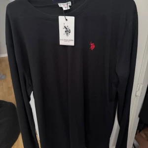 Svart långärmad tröja U.S. Polo Assn. - Svart långärmad tröja från U.S. Polo Assn. med klassisk rund halsringning och röd broderad logga på bröstet. Tröjan är i mjuk bomull och har en clean, stilren look som passar perfekt till jeans eller chinos. Fick i julklapp men den är köpt på zalando